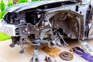 Front view of a car missing its bumper and front wheel, showing exposed mechanical parts and wiring. The car is propped on a jack stand, conveying a tone of repair or disassembly.