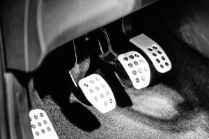 Close-up of three metal car pedals—clutch, brake, and accelerator—in black and white. The image highlights the textured surfaces and shadows.